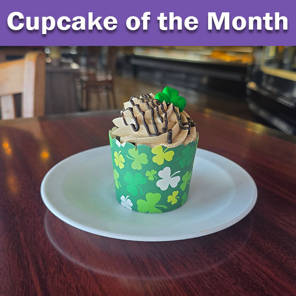 A chocolate cupcake in a green liner with shamrocks all over a mocha buttercream and a sugar shamrock on top with a chocolate ganache drizzled on top.